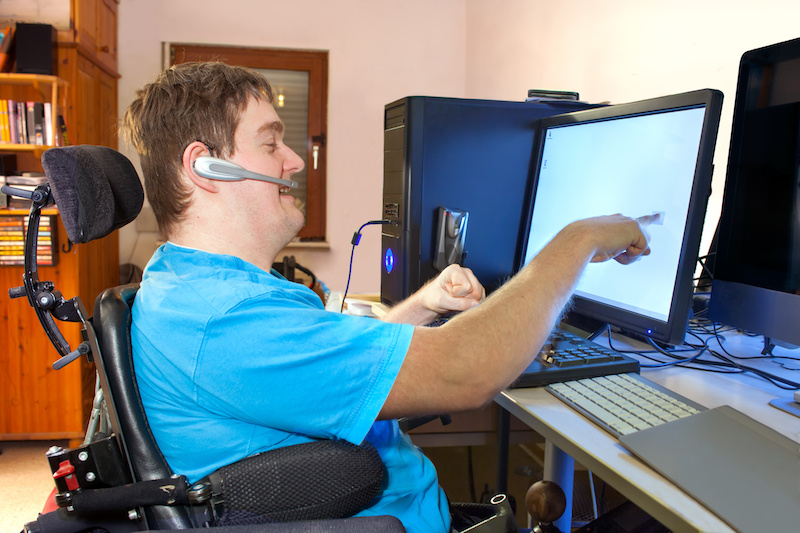 Spastic young man with infantile cerebral palsy caused by a complicated birth sitting in a multifunctional wheelchair, using a computer with a wireless headset, reaching out to touch the touch screen.